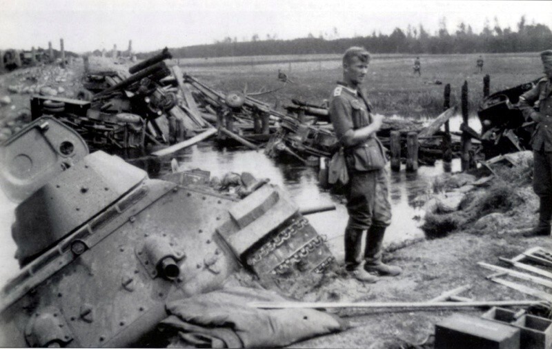 Немецкий фельдфебель возле советского танка Т-34 на разбитой переправе через речку Зельвянка. На переднем плане танк Т-34 образца 1941 года, в реке утопленный танк Т-34 образца 1940 года с пушкой Л-11. 