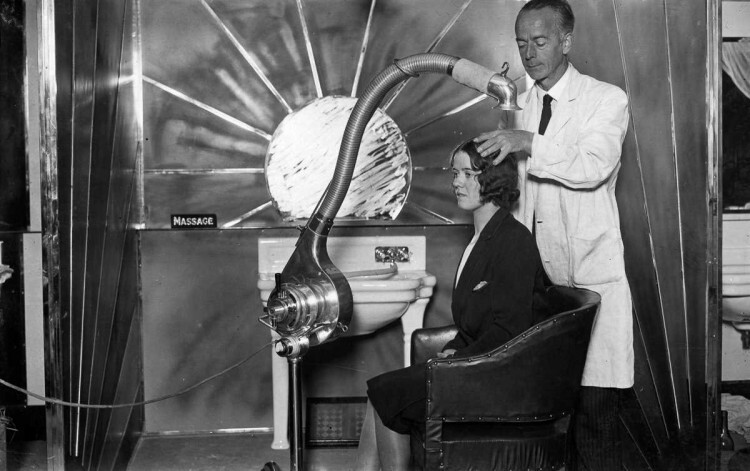 1929 г. Стилист использует автономный регулируемый фен для сушки волос клиентки в Лондоне.