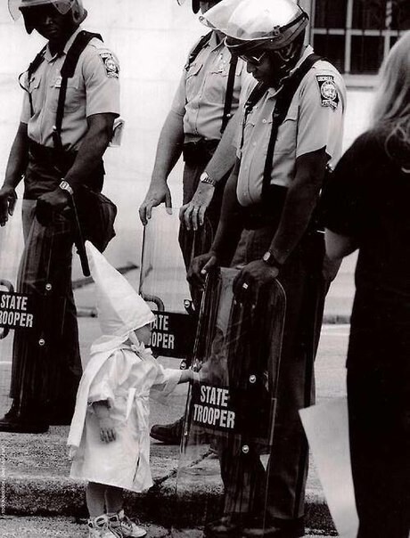 Ребенок члена организации Ку-клукс-клан, играющий со своим отражением на щите афро-американского полицейского во время демонстрации.