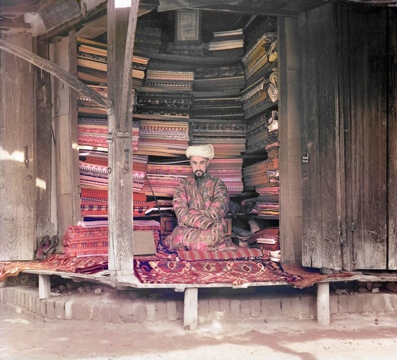 Купец в Самарканде торгует тканью из хлопка, шелка, шерсти и других материалов, коврами и другими товарами. Над тканями висит страница Корана