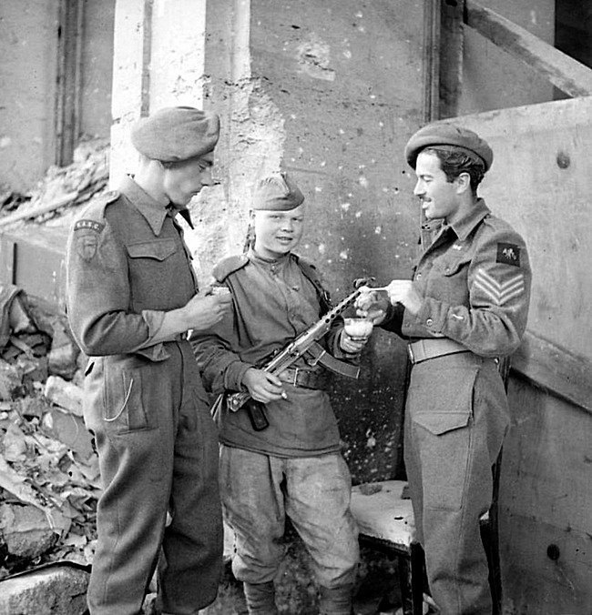 Британские сержант Р.С. Бейкер (R.S. Baker) из армейской кино-фотографического группы (AFPU) и солдат корпуса обслуживания (RASC), едят хлеб с солью с молодым советским солдатом у здания рейхсканцелярии в Берлине, в честь капитуляции немецких войск.