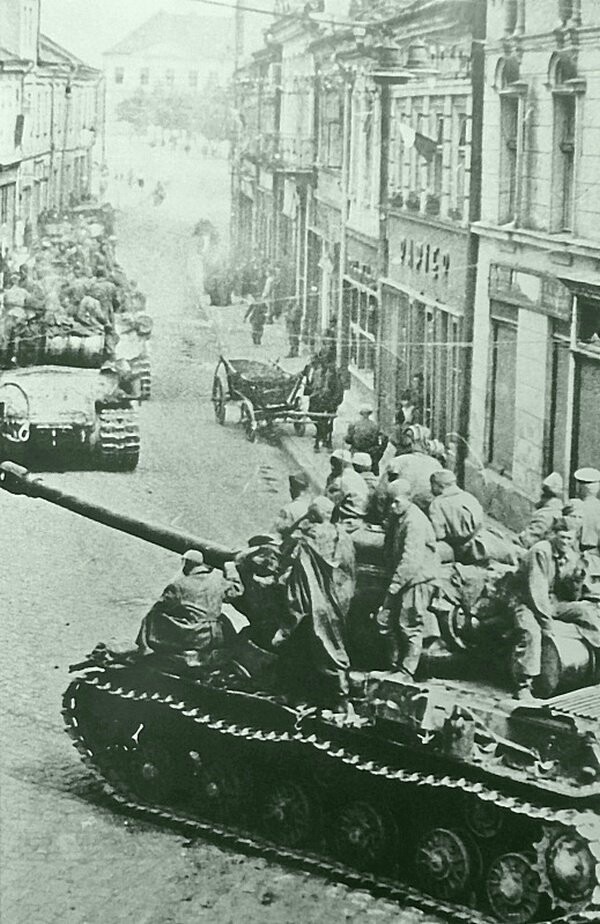 Колонна советских тяжелых танков ИС-2 с десантом на броне на улице взятого города в восточной Германии.  На одном из фасадов справа виден флаг, напоминающий польский (бело-красный); возможно, что  - вопреки описанию в источнике - фотография сделана н
