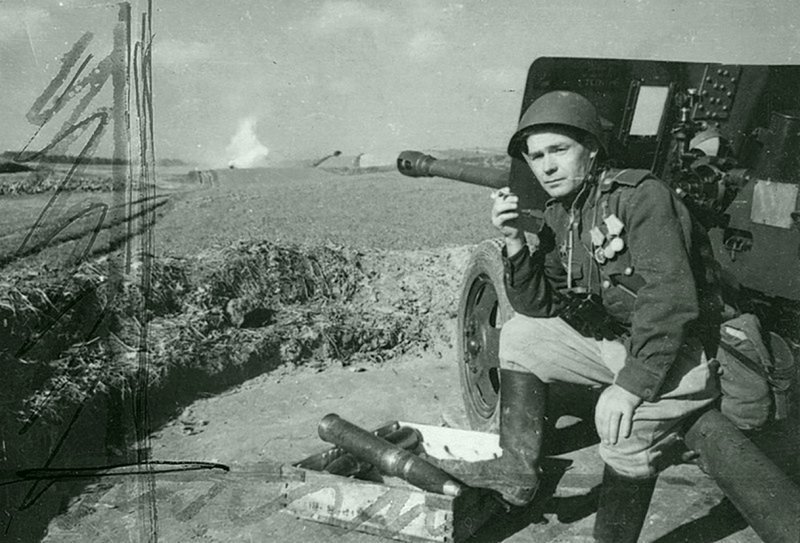 Герой Советского Союза ефрейтор-артиллерист курит, сидя на станине 76,2-мм дивизионного орудия ЗИС-3 образца 1942 г.  Автор: Дмитрий Бальтерманц.