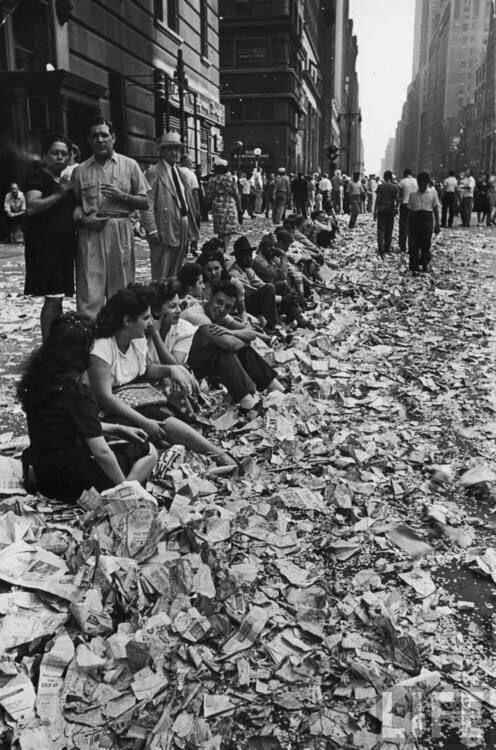 Люди, сидящие на бордюре среди конфетти и бумаг после празднования окончания Второй мировой войны в Нью - Йорке 14 августа 1945 года