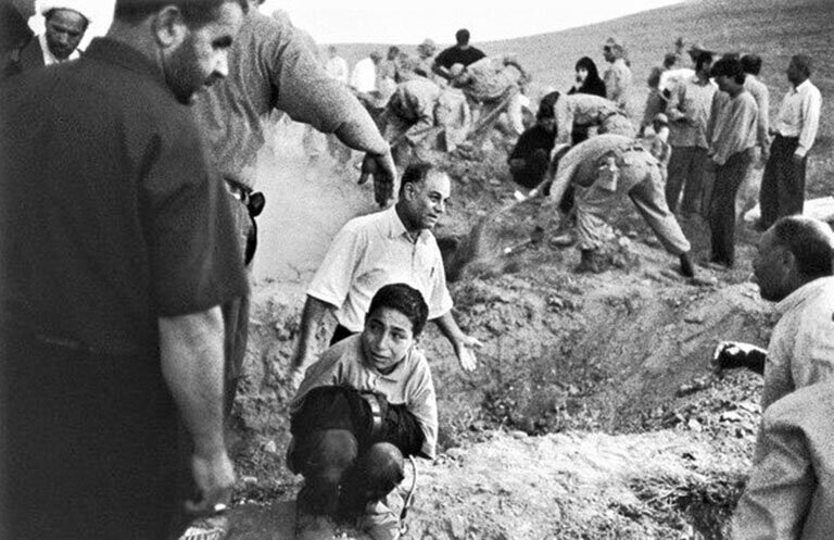 Окруженный солдатами и жителями, роющими могилы для жертв землетрясения, мальчик держит брюки своего мертвого отца и сидит на корточках около места, где его отец будет похоронен. 23 июня 2002 года. Провинция Казвин, Иран. 