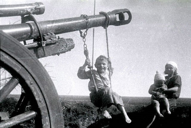 Мальчик качается на качелях, подвешенных на дуло брошенного немцами орудия. Рядом девочка с маленьким ребенком на коленях. 1944 г. Украина, Николаевская область.