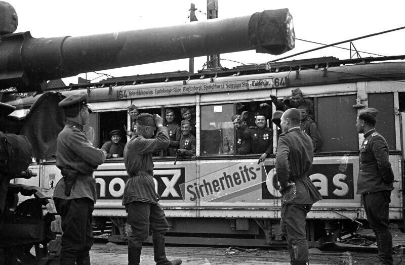 Советские бойцы и командиры в вагоне берлинского трамвая. Их «провожают» товарищи, машут вслед. Слева в кадре виден ствол 203-мм гаубицы Б-4. 1945