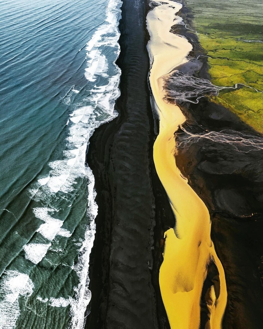 Синее море накатывается на черный пляж, по которому течет желтая из-за серы река, обрамленная зелеными полями
