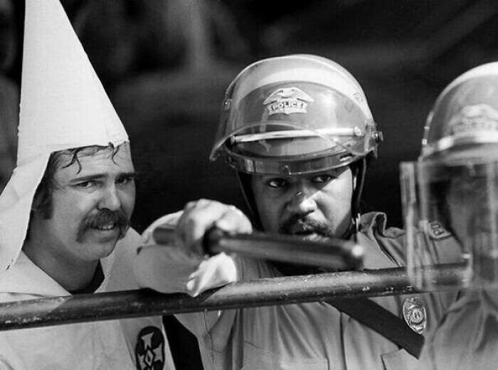 Член Ку-клукс-клана стоит за полицейским для защиты после того, как толпа окружила его митинг его клана в Остине, Техас, 1983.