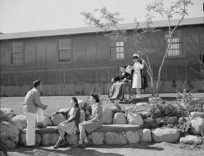 16. Медсестра стоит за мужчиной в инвалидной коляске, пока на переднем плане молодой человек разговаривает с двумя девушками.