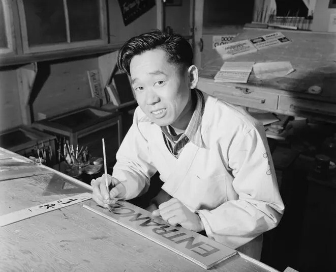 23. Акио Мацумото рисует вывеску, сидя за столом.