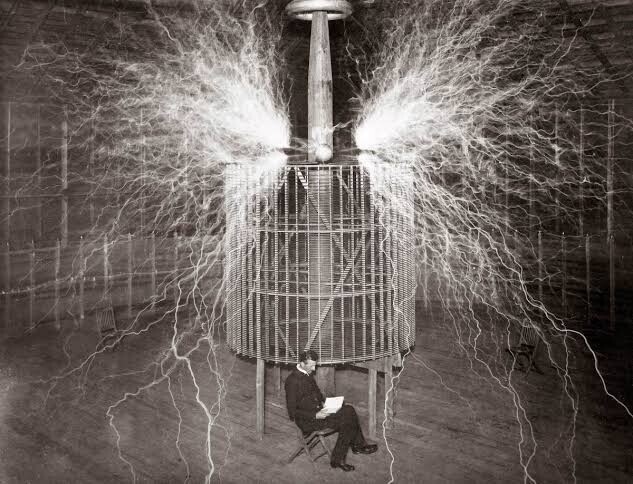 Никола Тесла сидит в своей лаборатории в Колорадо-Спрингс рядом со своим увеличительным передатчиком высоковольтного генератора, в то время как он производит электрические разряды. Декабрь 1899 год