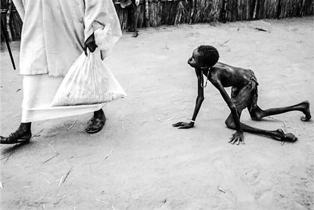 Изнеможённый от голода мальчик ползёт за взрослым с мешком еды. Судан, 1998 год