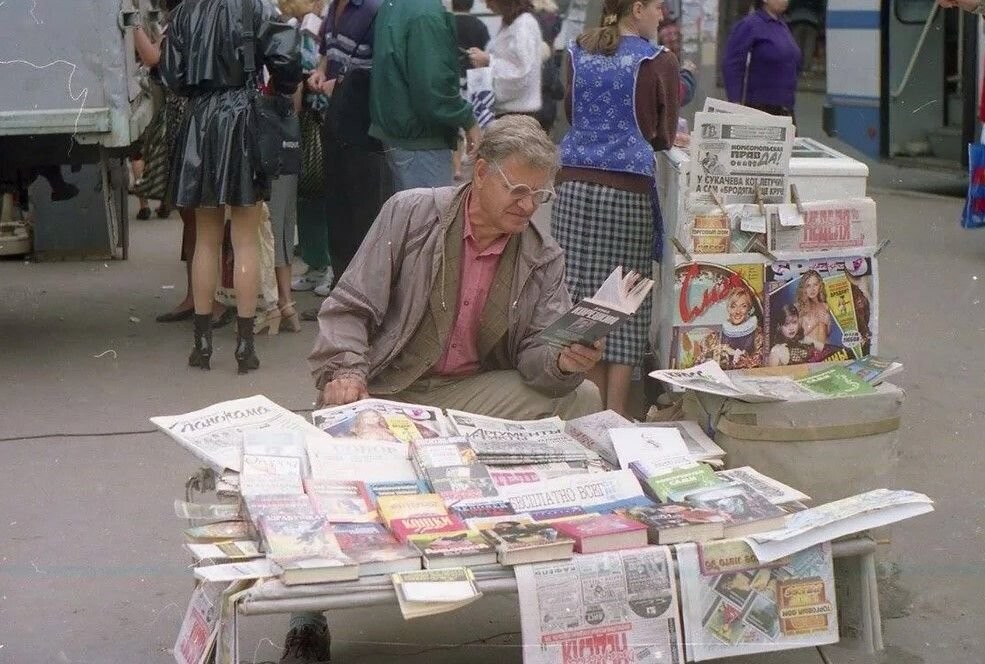 Мужчина продаёт печатную продукцию, попутно читая книгу. Россия, 1990-е годы.